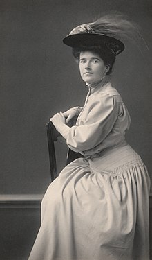A young white woman with dark hair in a bouffant updo, wearing a hat and seated in a chair