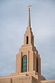 The spire of the Red Cliffs Utah Temple dominates the view of a portrait shot. A multilevel spire has rounded windows on the sides, along with a golden statue at the top of the angel Moroni, against a backdrop of a blue sky.