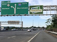 A multilane freeway approaching an interchange with a sign display over the road. The left sign reads exit 66 south Route 17 Mahwah left two lanes exit Interstate 287 south Morristown right two lanes straight and the right sign says Welcome to New Jersey
