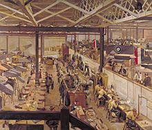 interior view from an elevated position showing the shop-floor of an aircraft assembly factory. The aircraft are seen in various stages of assembly to the left and right of the image. In the centre are lines of workbenches and a range of different factory personnel