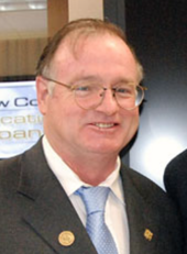Color photograph of a white man in glasses wearing a collared shirt, tie, and jacket