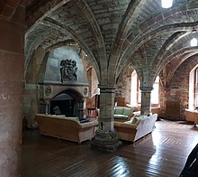 Undercroft of Newbattle Abbey College. It has an empty fireplace to the left of the image, with sofas in front. Stone arches gracefully support the ceiling above.