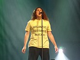A man standing behind a microphone stand, wearing a yellow shirt that contains the text „Atlantic Records Sucks“