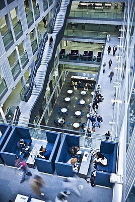 The image shows the inside of the Manchester Metropolitan University Business School. The image is taken from a corner, showing several floors within the building. You can clearly see staff and students walk around the building.