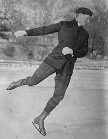 Man in his 30s, wearing dark trousers and coat, skating on an outdoor rink, facing to the right and looking downward towards the ice