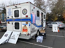 An ambulance parked in a parking lot that has been turned into a mobile COVID testing center is festooned with a trans pride flag and pro-LGBT messages. A sign next to it says "testing center."