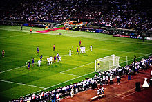 A wide-angle photograph of several football players preparing for a free-kick to be taken.