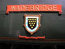 A 'West Country' class enamelled metal nameplate and shield mounted on flat metal casing covering the locomotive boiler. The nameplate comprises a scroll, and below this is a shield containing a picture of a coat-of-arms. A second scroll is below the shield, allowing identification as a member of the 'West Country class'.