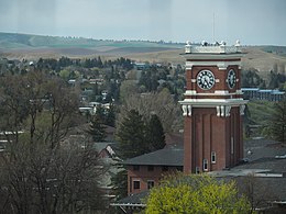 A brick clock tower with white clock faces and black Roman Numerals in front of rolling fields.