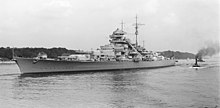 A large light gray warship bristling with guns sits in harbor