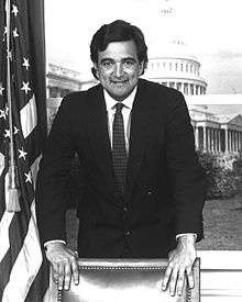 A Latino man leans over a chair and smiles at the camera, with the U.S. Capitol in the background