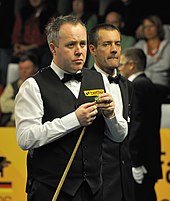 John Higgins stands holding his cue in his right hand while he chalks it with his left hand.