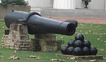 A long 19th-century cannon with a stack of cannonballs in front of it, sitting on a lush green lawn.