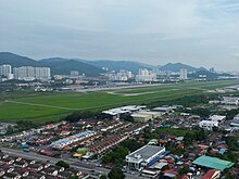 Aerial view of an airport, with factories at the foreground. More buildings and hills form the backdrop.