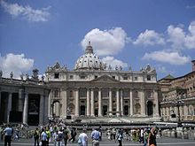 Frontale Farbfotografie vom Petersdom mit dem Platz und vielen Menschen im Vordergrund. Ein Teil des linken Säulengangs vom Platz ist zu sehen. Die Säulen verlaufen über zwei Geschosse. Auf dem Dach sind Marmorfiguren und eine Kuppel.