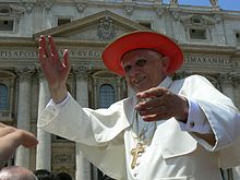 Farbige Untersicht vom Papst in weißem Papstgewand mit einem goldenen Kreuz und einem roten breitkrempigen Hut. Er streckt seine Arme zur Begrüßung aus. Im Hintergrund ist ein altes Gebäude zu sehen.