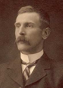 Head and shoulders portrait of a man with a moustache in a suit and tie with a high collar.