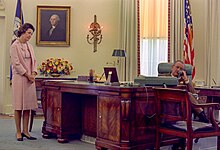 Johnson sitting behind the desk on the phone while Lady Bird Johnson stands to the side dressed in pink.