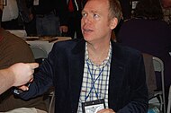 A man with the shot focused on his upper body. He is wearing a dark colored jacket and a white and gray shirt.
