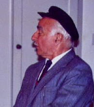 a colour photograph of the left profile of a gray-haired man wearing a blue suit, a tie and a flat cap