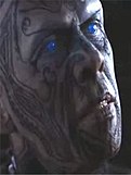 A closeup of a man's head; his face is covered in tattoos, and his eyes are glowing blue.