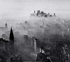 A black-and-white, panoramic view of New York City as seen from a great height. A vast number of buildings and skyscrapers can be seen. A hazy, smoky gas overlays the entire city like a blanket, with a fairly clear skyline only in the far distance at the horizon. Near the closest buildings, the smog appears thin and wispy. The smog appears thicker and thicker around buildings that are farther away from the photographer's position, until shorter buildings near the horizon are almost entirely shrouded and impossible to see under a thick layer of smog. Near the horizon, the clustered tops of tall skyscrapers emerge from within the smog.