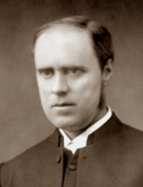 Bald white man in middle age, wearing clerical collar and black clothing