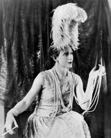Young white woman in elaborate 1920s evening costume, with copious pearl necklace and holding a cigarette in a long holder