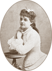 teenage white woman in 19th-century costume seated, with both elbows on a side table and her hands to her face