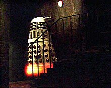 In a dark basement, a white Dalek (see previous description) appears to levitate up a small staircase of approximately seven stairs. The body of the Dalek is white, with shiny gold vertical slats and gold balls on its lower half. There is an orange-yellow glow at the Dalek's base.