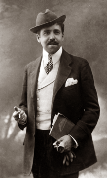 photograph of elegantly dressed white man with neat moustache and beard, wearing a hat at a jaunty angle; he has a cigarette in his right hand