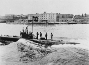 A surfaced submarine moves forward with its crew standing on the deck and conning tower. The city of Pola can be seen in the background and the naval ensign of Austria-Hungary flies from the submarine's conning tower. The main entry hatch of the boat is open with a submariner standing half way inside.