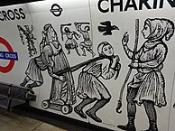 A platform wall: decorative panels show wood-cut images of people in mediaeval dress. Two push and pull a statue on a wheeled trolley as another beckons them forward. Two men in the background fight with clubs and another sits on a tower reading