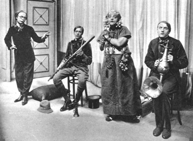 Stage group of four middle-aged white people in extravagant clothing; two of the men hold ornate and improbable musical instruments and the one woman is declaiming