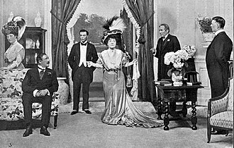 stage scene with grand dame centre, in large Edwardian hat, with men in evening clothes to her left and right