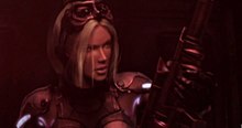 A young blonde woman in an armored suit loads a sniper rifle in a red lit room.