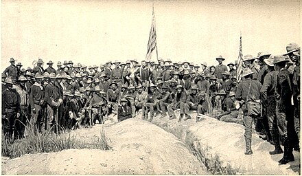 A black-and-white photo of US Army soldiers on July 3, 1898, in an upside-down V-type formation on top of Kettle Hill, two American flags in center and right. Soldiers facing camera.