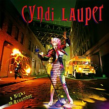Cyndi Lauper (seen wearing a colorful outfit) holding a microphone that is attached to a stand on a cobblestone street. The Stand is seen nearly leaning while Lauper is holding the stand. The Manhattan Bridge can be seen in the background. Near Lauper, a School Bus and another person doing a fire performance on the sidewalk.