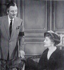 Middle-aged man with pencil moustache standing, with knowing expression, talking to seated middle-aged woman. She is wearing a hat, and he wears a black mourning armband.