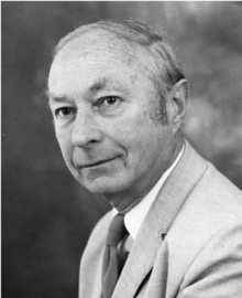 Black-and-white photograph of an elderly white man, wearing a grey suit and dark tie, his hair combed over to the right.
