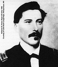 Black-and-white picture of a white male with a full dark mustache and squared goatee facing the camera and looking slightly to the right: He has dark, swarthy hair neatly combed over a broad forehead. His dress uniform has a white collar with the front ends turned down with a small bow tie.