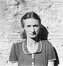 A woman with shoulder-length hair, photographed against a wall or cliff face.