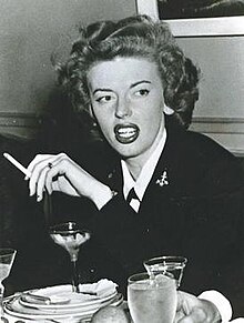 A young white woman in a black blazer. She is holding a cigarette and there is a martini glass on a table in front of her.