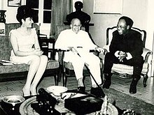 An elderly, smiling Du Bois sits in a chair, flanked by a man and woman also seated and smiling