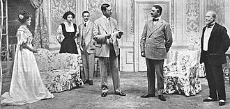 scene from a stage play with six characters – two women and three men – in Edwardian dress grouped in a luxurious drawing room