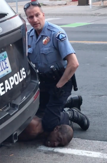 Two men on an asphalt surface, behind a black van on which the letters "EAPOLIS" is seen, with a license plate ending "ICE". One man has light skin, a blue shirt with identifying badges on his chest and shoulder, black pants and boots, and black sunglasses pushed to the top of his close-shorn head. He is kneeling with his left knee and upper shin resting on the neck of the other man, and his right knee out of sight behind the van. The other man is lying prone, with his left cheek pressed against the asphalt close to a painted line. He is dark-skinned, with similarly short hair, and is not wearing a shirt; His mouth is slightly open, his eyes are closed with his eyebrows raised, and his arms are down, not visible behind the van. The kneeling man has his left hand in a dark glove, with his right arm hidden behind the van, and is looking at the viewer with his eyebrows slightly lifted and mouth slightly open.
