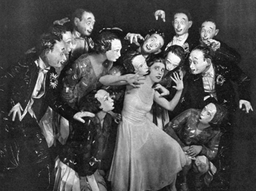 young white woman in white evening dress surrounded by menacing figures in evening clothes and scary face masks