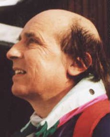 Simon Clarke looking towards the top-left corner with his mouth slightly open. The top of his head is bald, but he has brown hair elsewhere. He is wearing a colorful purple, red, and green shirt with a white collar.
