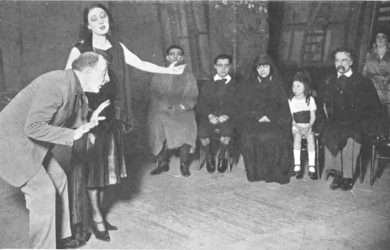 stage scene with director bending forwards, actress standing gesticulating and small family sitting in row