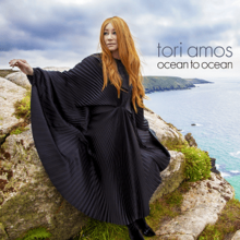 A photo of the artist wearing a black cloak and stood toward the edge of a cliff overlooking the ocean.
